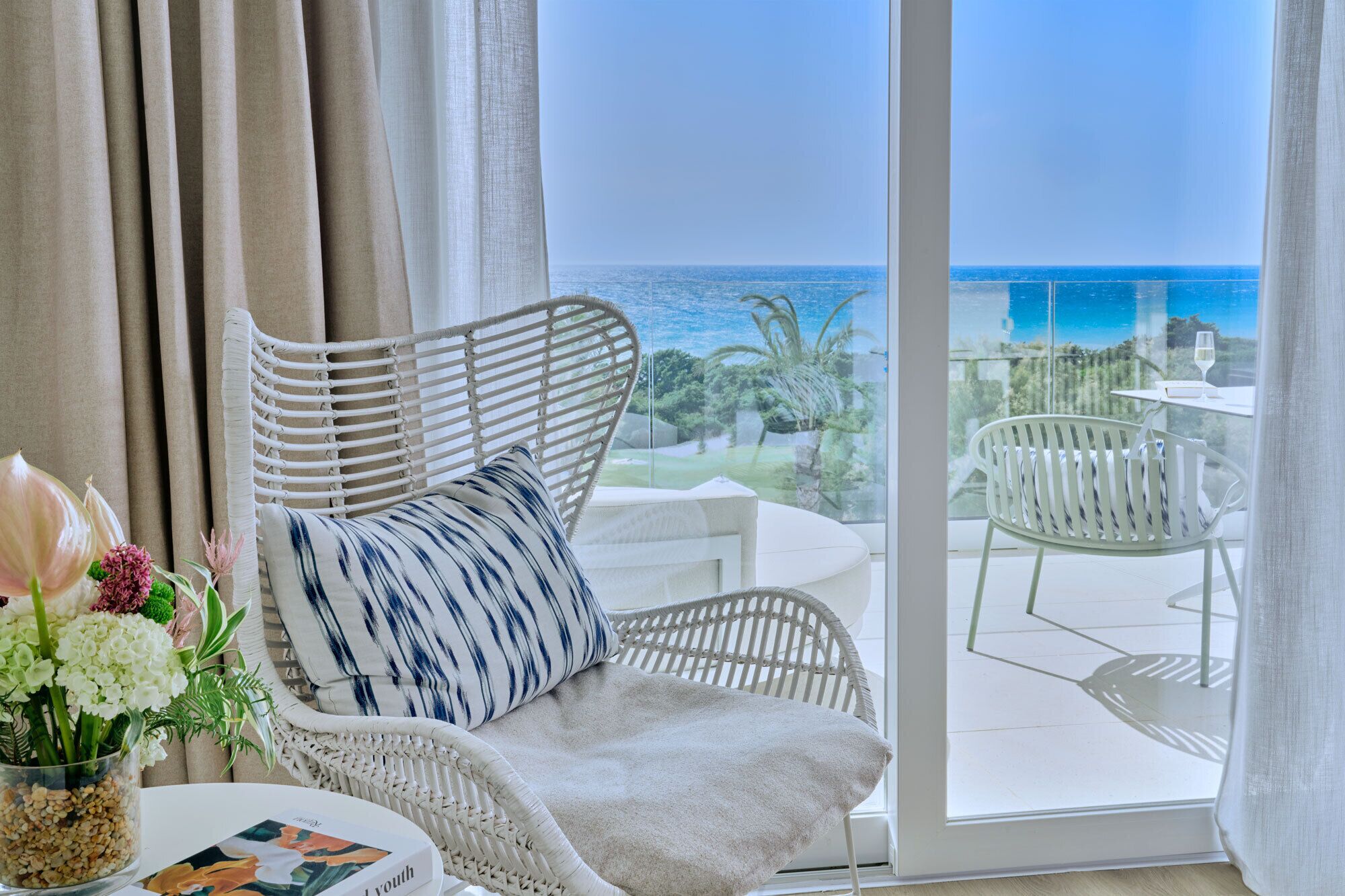 A room with a view of the ocean, a wicker chair, a table with a glass, and a vase with flowers.