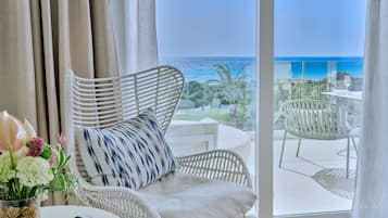 A room with a view of the ocean, a wicker chair, a table with a glass, and a vase with flowers.