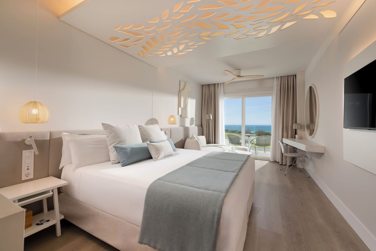 Quarto de hotel moderno com cama grande, uma mesa, televisão e vista para o oceano.