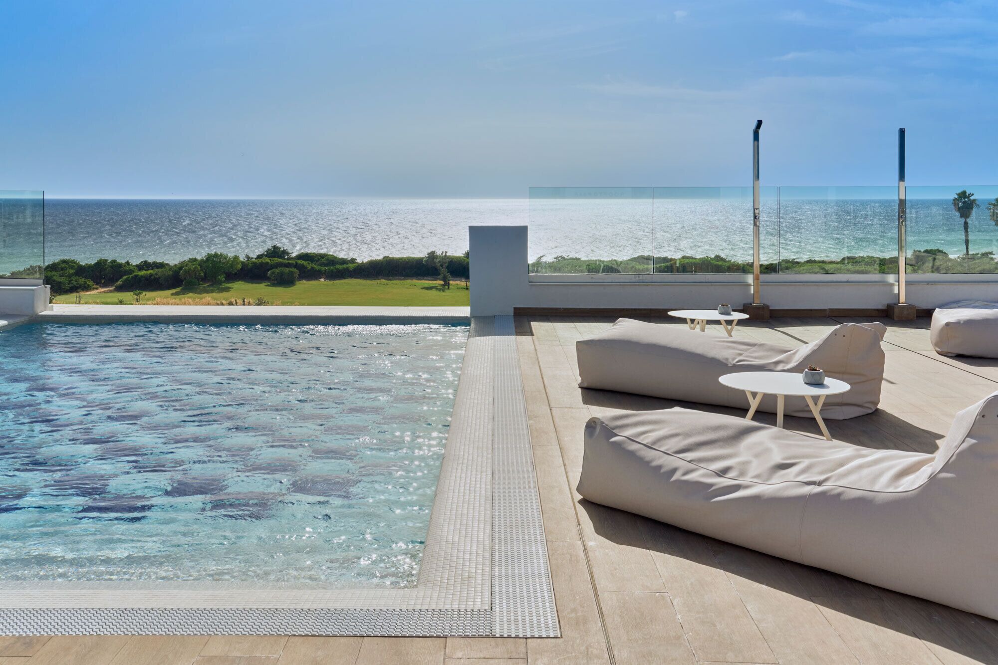 A rooftop pool area with a clear view of the ocean, lounge seating, and a glass balustrade.