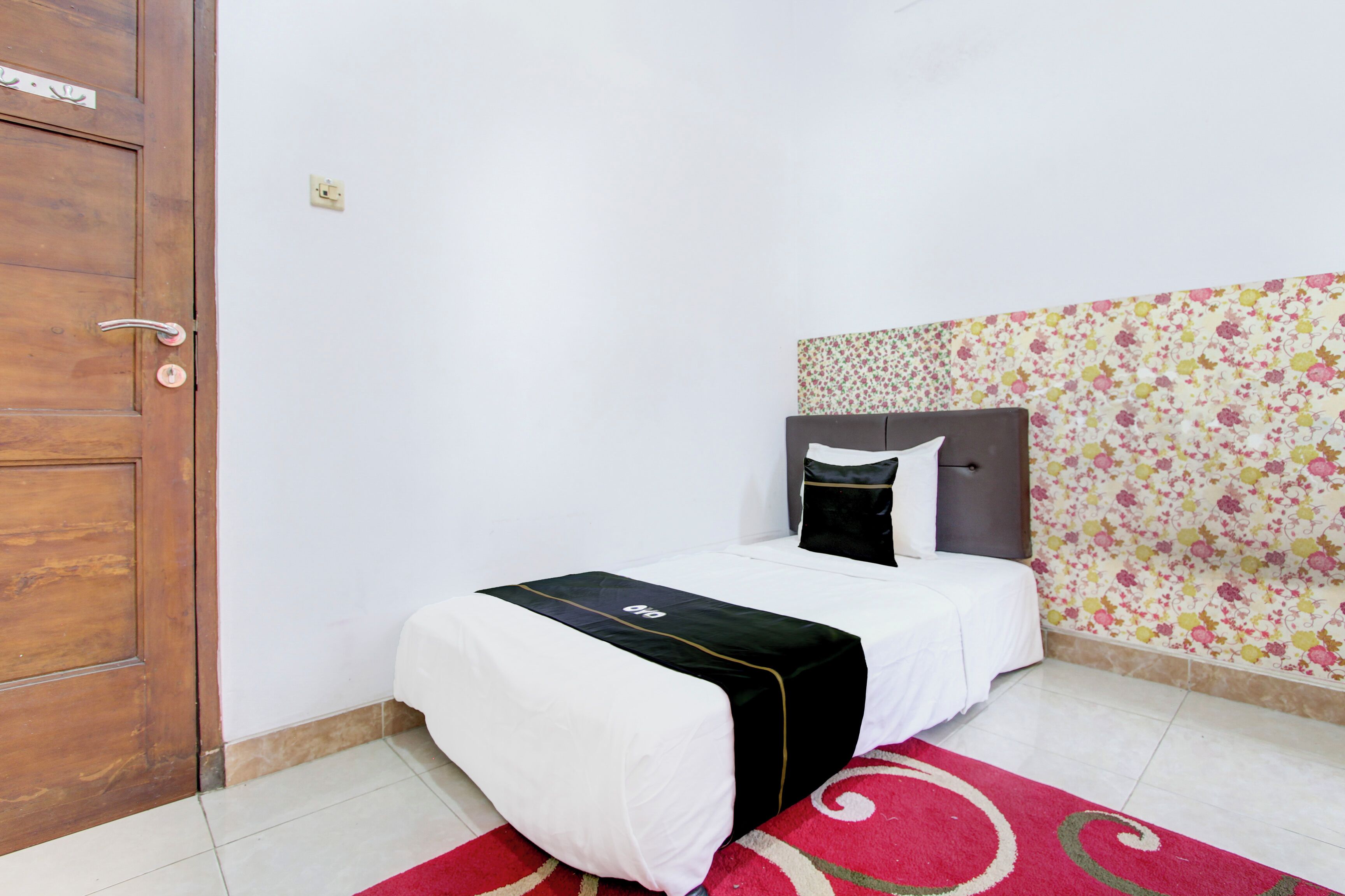 Una cama bien tendida con una colcha blanco y negra, un tapete rojo y beige, y una pared con estampado floral.