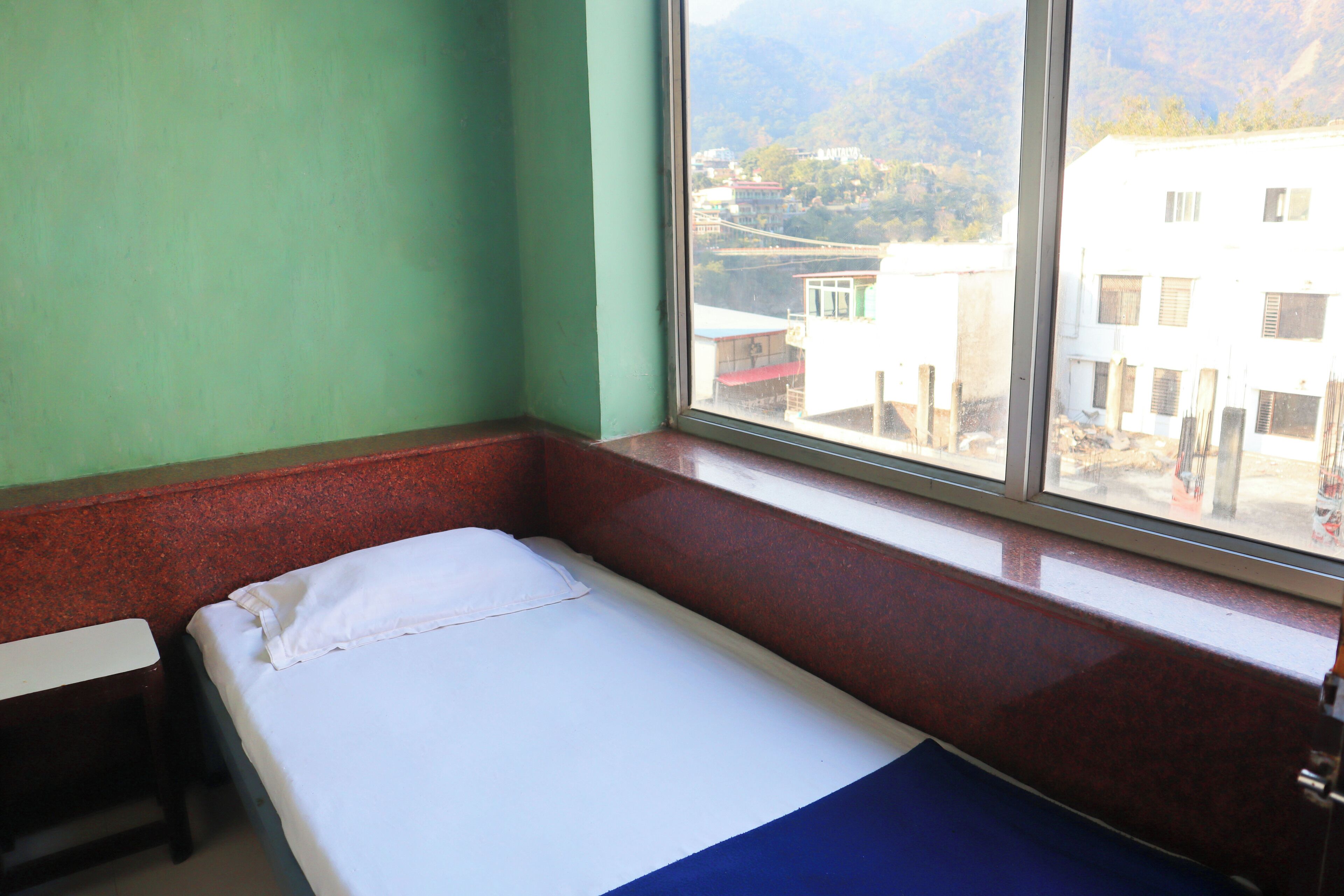 Una cama individual con colchón blanco y azul, una mesita de noche de madera y una ventana con vistas a edificios y montañas.