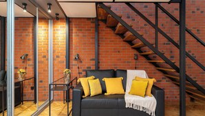 Un salon moderne avec un canapé en cuir noir, des coussins jaunes, un escalier en bois et un mur en briques.