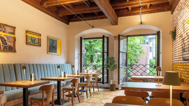 Un restaurant avec des tables et des chaises en bois, un banc bleu rembourré et de grandes fenêtres donnant sur un paysage verdoyant.