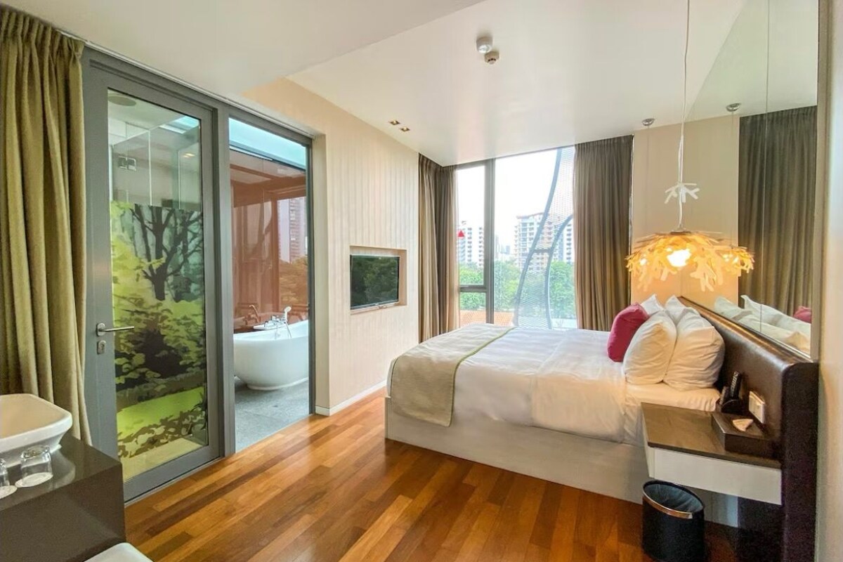 Quarto de hotel moderno com uma cama grande, banheiro com banheira, porta de vidro deslizante para uma varanda e vista para a cidade.