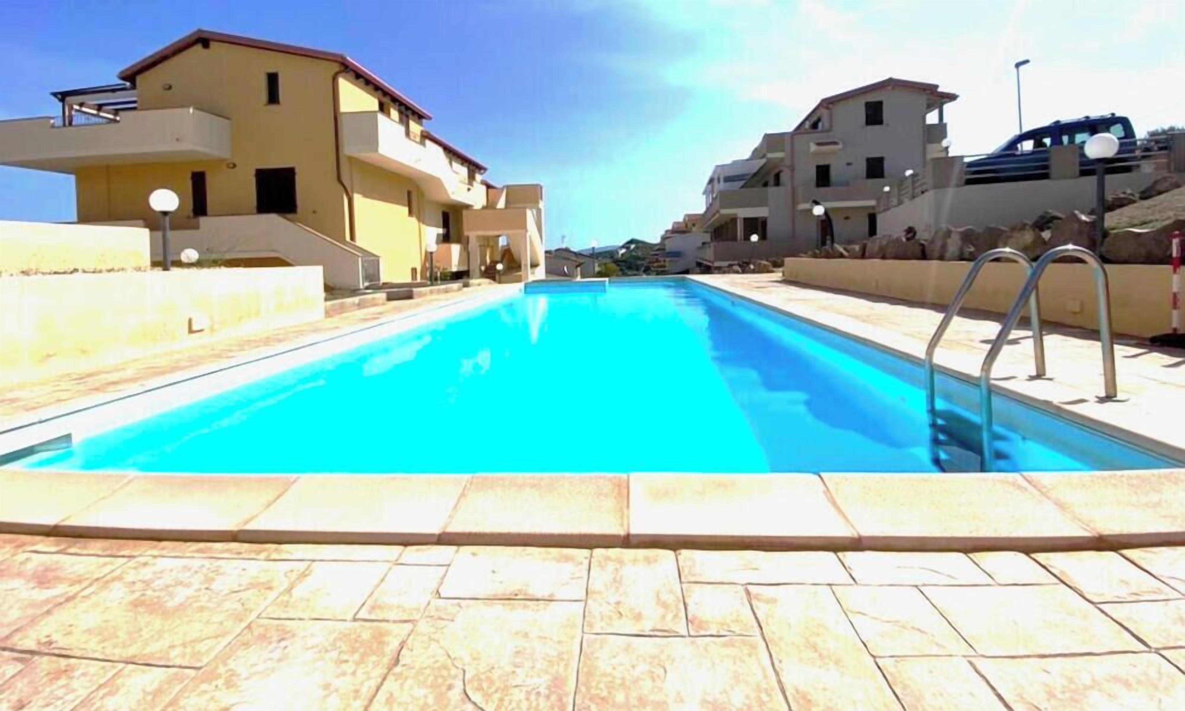 A swimming pool with a tiled deck, surrounded by buildings with balconies and a clear sky.