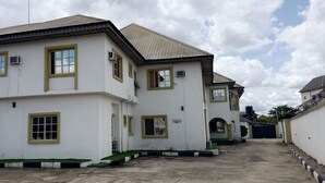 A white building with yellow trim and multiple windows, a paved entrance, and a gated driveway. - Calawazobia hotel (Uyo)