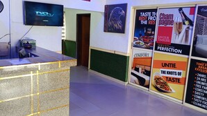A hotel reception area with a counter, a television displaying 'DSLV', and various posters advertising the hotel's amenities and services. - Calawazobia hotel (Uyo)