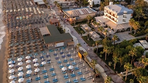 An aerial view of a beach resort with rows of sunbeds and umbrellas, a central building with a roof, and surrounding palm trees. - Vila Lasko (Golem)