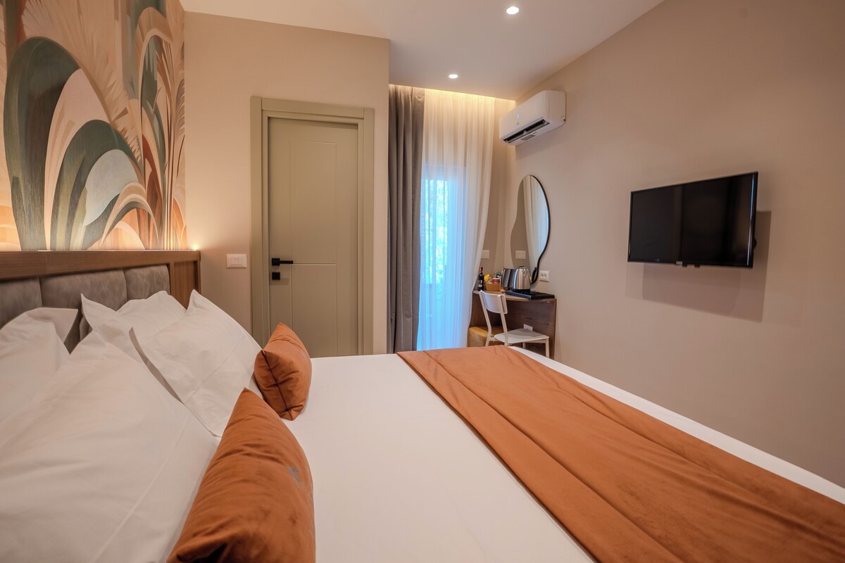 A hotel room with a large bed, a desk with a chair, a flat-screen TV, and an air conditioning unit on the wall.