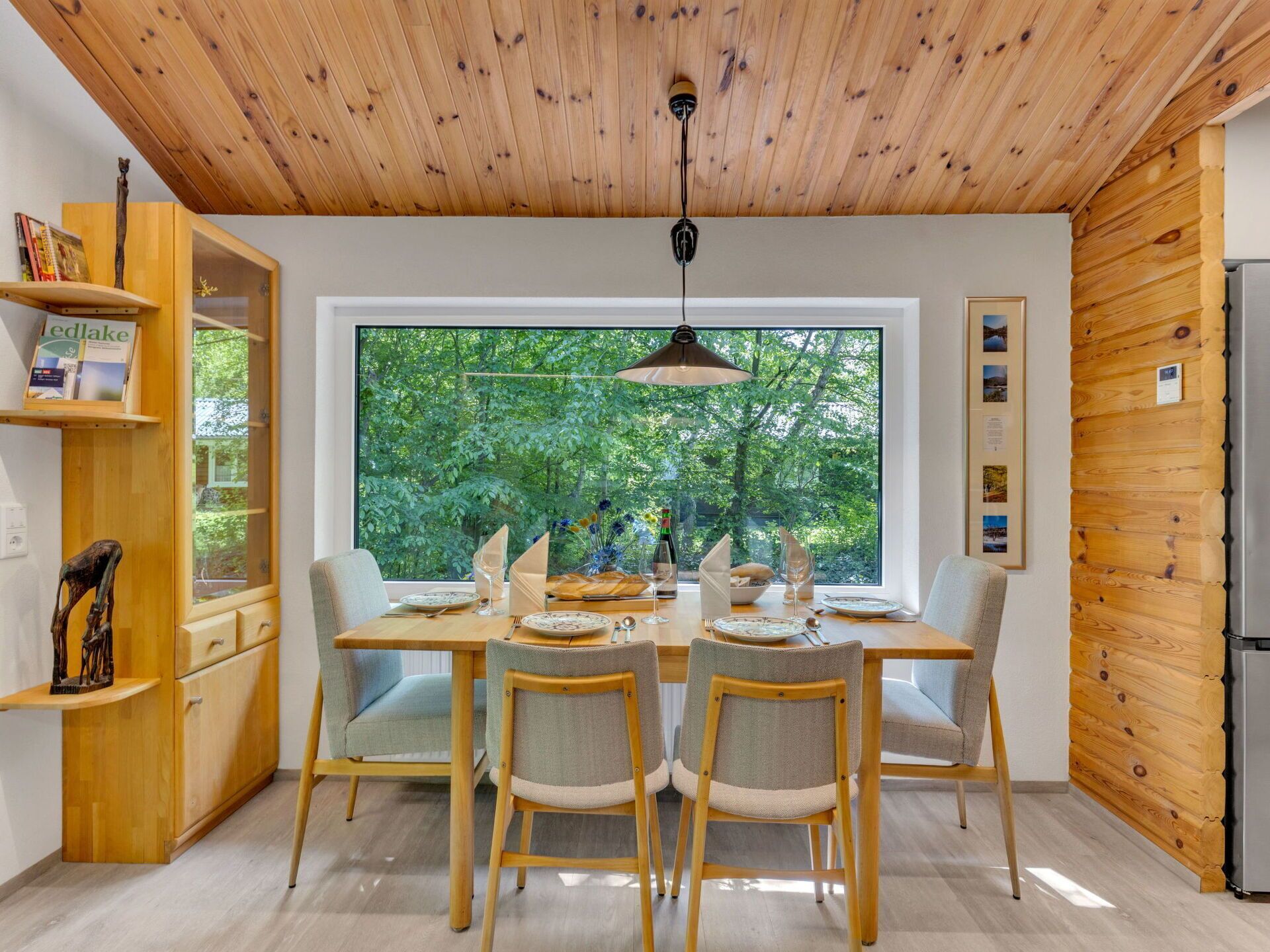 Een moderne keuken met houten plafond en wanden, een eettafel voor vier personen en een groot raam met uitzicht op bomen.