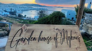 Una persona sostiene un letrero de madera con el texto 'Garden house of Mykonos'.