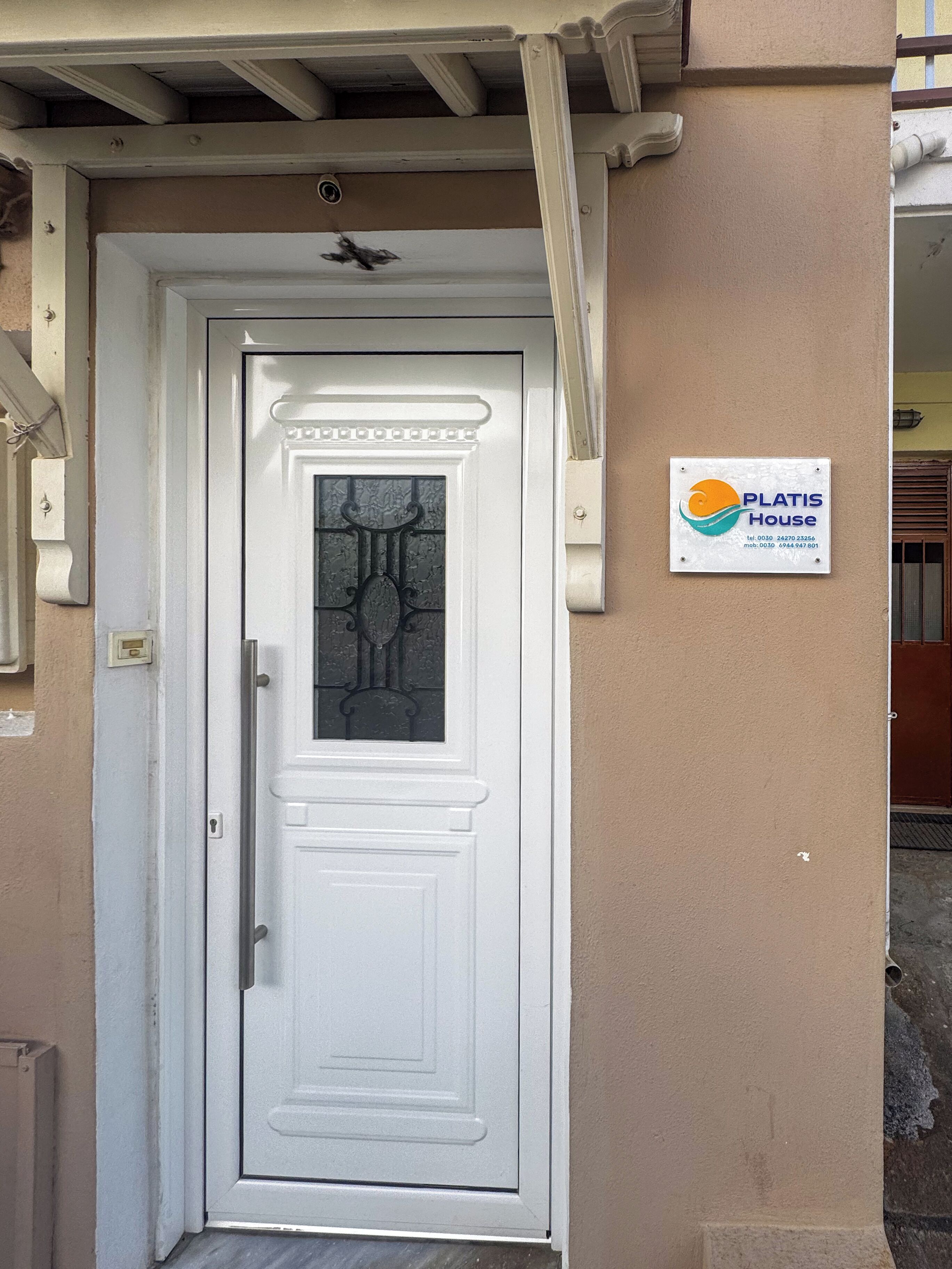 A white door with a glass panel and a decorative top. A sign with the text "PLATIS House" is mounted on the wall to the right of the door.