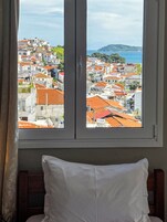 Una habitación con cama, ventana y vistas a un pueblo costero con tejados rojos.