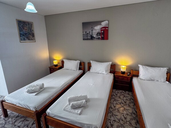 A hotel room with three single beds, each with a wooden headboard and white bedding. There are bedside tables with lamps and a painting of a cityscape on the wall. - Skiathos Platis House (Skiathos)