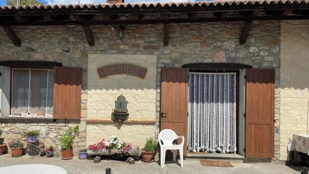 Un bâtiment en pierre avec un toit en tuiles, des volets en bois et un petit porche agrémenté de plantes en pot et d’une chaise en plastique blanche.