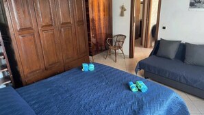 Une chambre avec deux lits, une armoire en bois, une chaise en osier et un canapé gris.