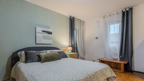 A bedroom with a bed, a bedside table, a lamp, a bench, and a window with curtains.