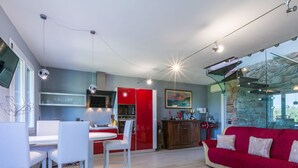 Una cucina moderna con armadietti rossi, un'area pranzo con sedie bianche e un divano rosso in un soggiorno con parete in pietra e ampie finestre.
