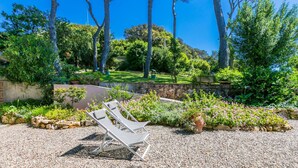 Ein Garten mit drei weißen Liegestühlen auf Kies, umgeben von üppigem Grün und blühenden Pflanzen.