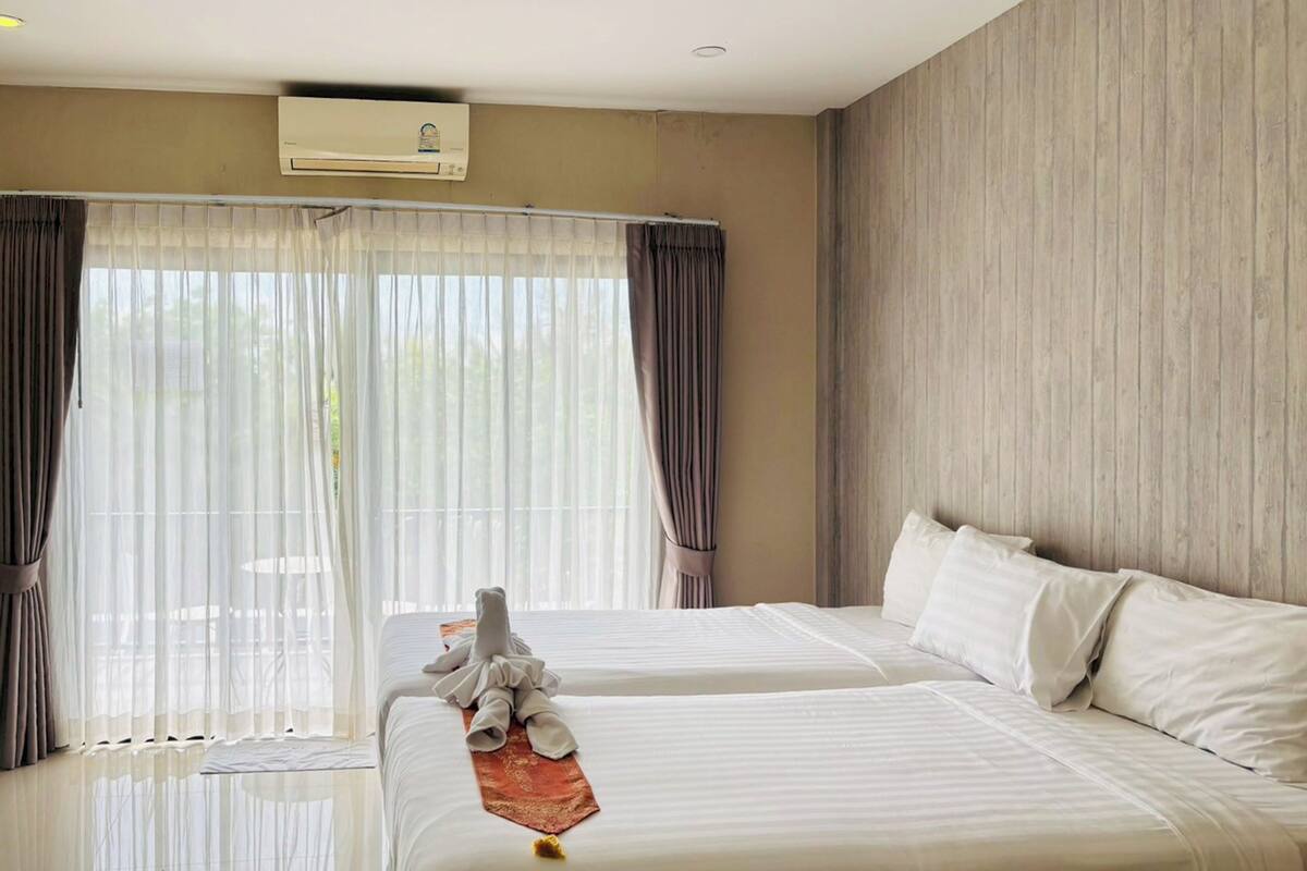 A hotel room with two beds, a large window with sheer curtains, an air conditioning unit, and a wooden wall.
