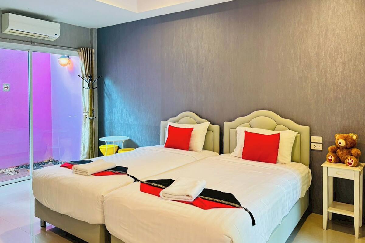 A double bed with white and red bedding, a small bedside table with a teddy bear, and a window with curtains.
