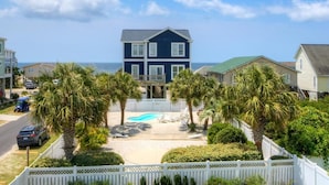 A two-story house with a swimming pool, surrounded by palm trees and a white picket fence.