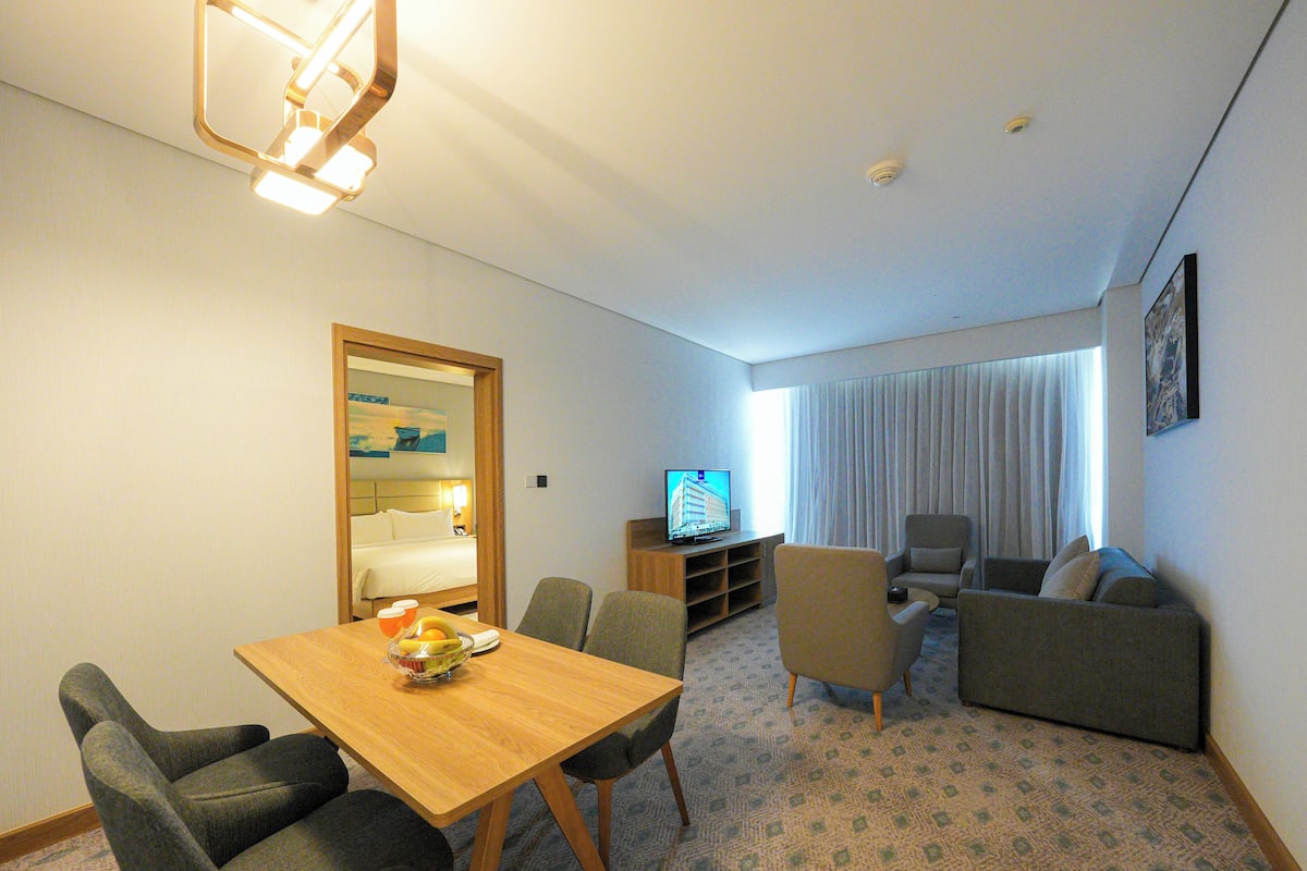 A hotel room with a dining area, a living room with a sofa and armchairs, a television, and a window with curtains.