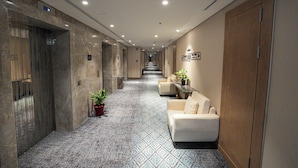 A hotel hallway with marble walls, a patterned carpet, a reception desk with a plant, and a wooden door on the right. - Casa Di Ora Hotel Al Rawdah (Jeddah)