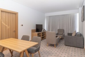 Un salon moderne avec une table en bois, des fauteuils gris, un canapé gris, un téléviseur et une grande fenêtre avec des rideaux.