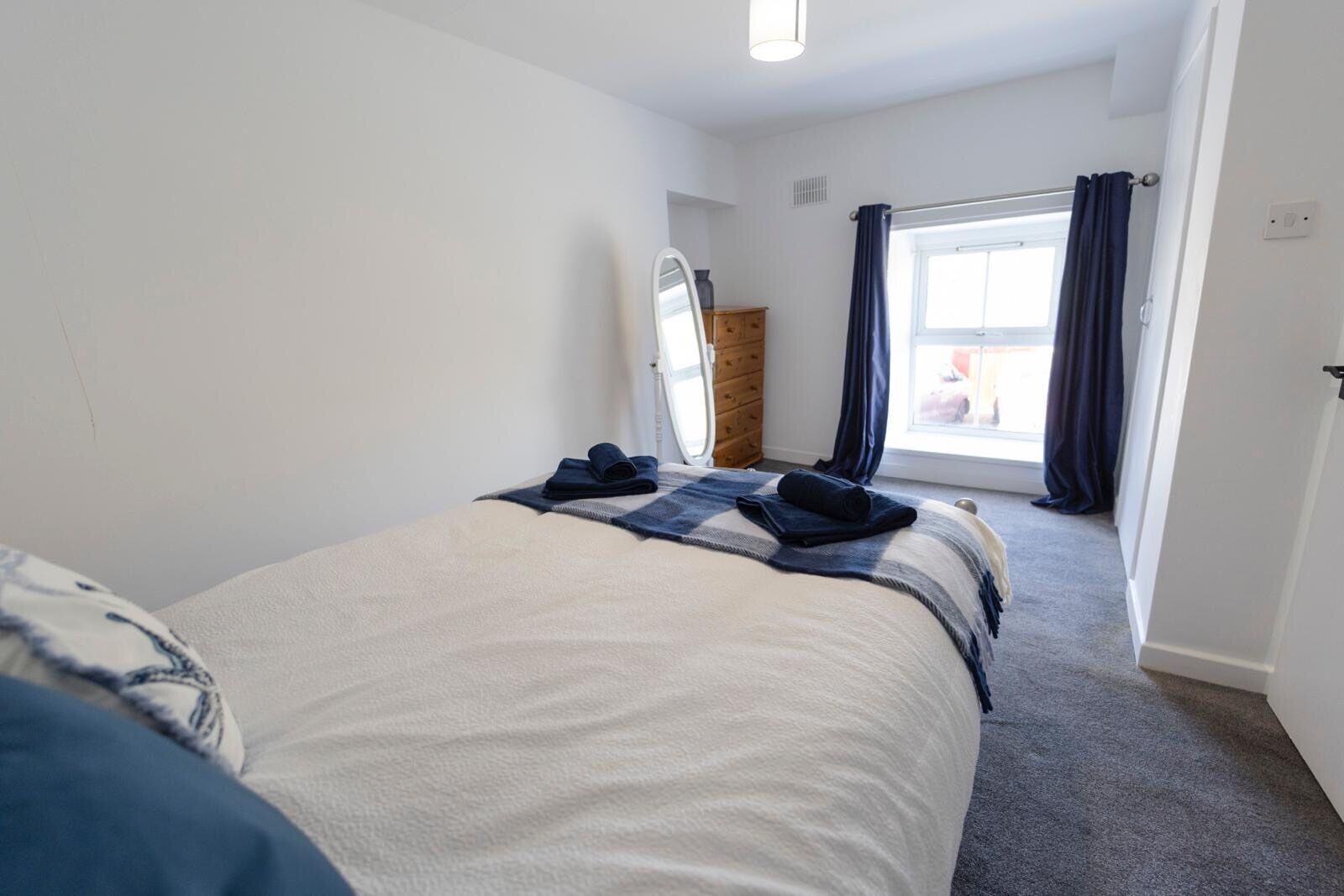 A bedroom with a bed, a window with curtains, a wooden door, and a small table with a mirror.