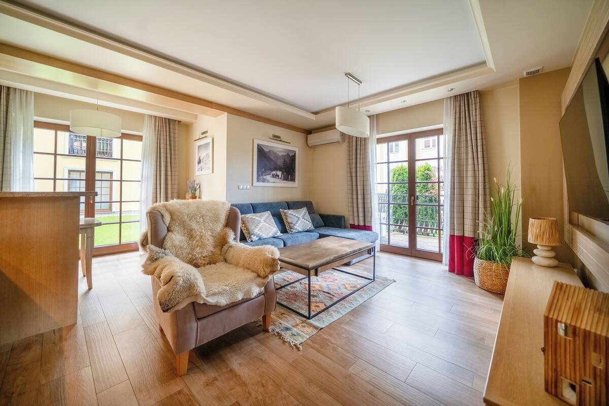 Amplia sala de estar con sofá, sillón y mesa de centro. Se aprecia un ventanal con cortinas, un aire acondicionado instalado en la pared y un televisor sobre un mueble de madera.