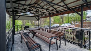 Un patio techado al aire libre con mesas de picnic y bancos, rodeado por una cerca de metal y árboles.