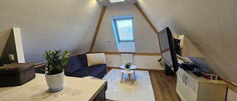 Un acogedor espacio tipo loft con un sofá azul oscuro, una mesita blanca, una planta en maceta y un televisor sobre un mueble.