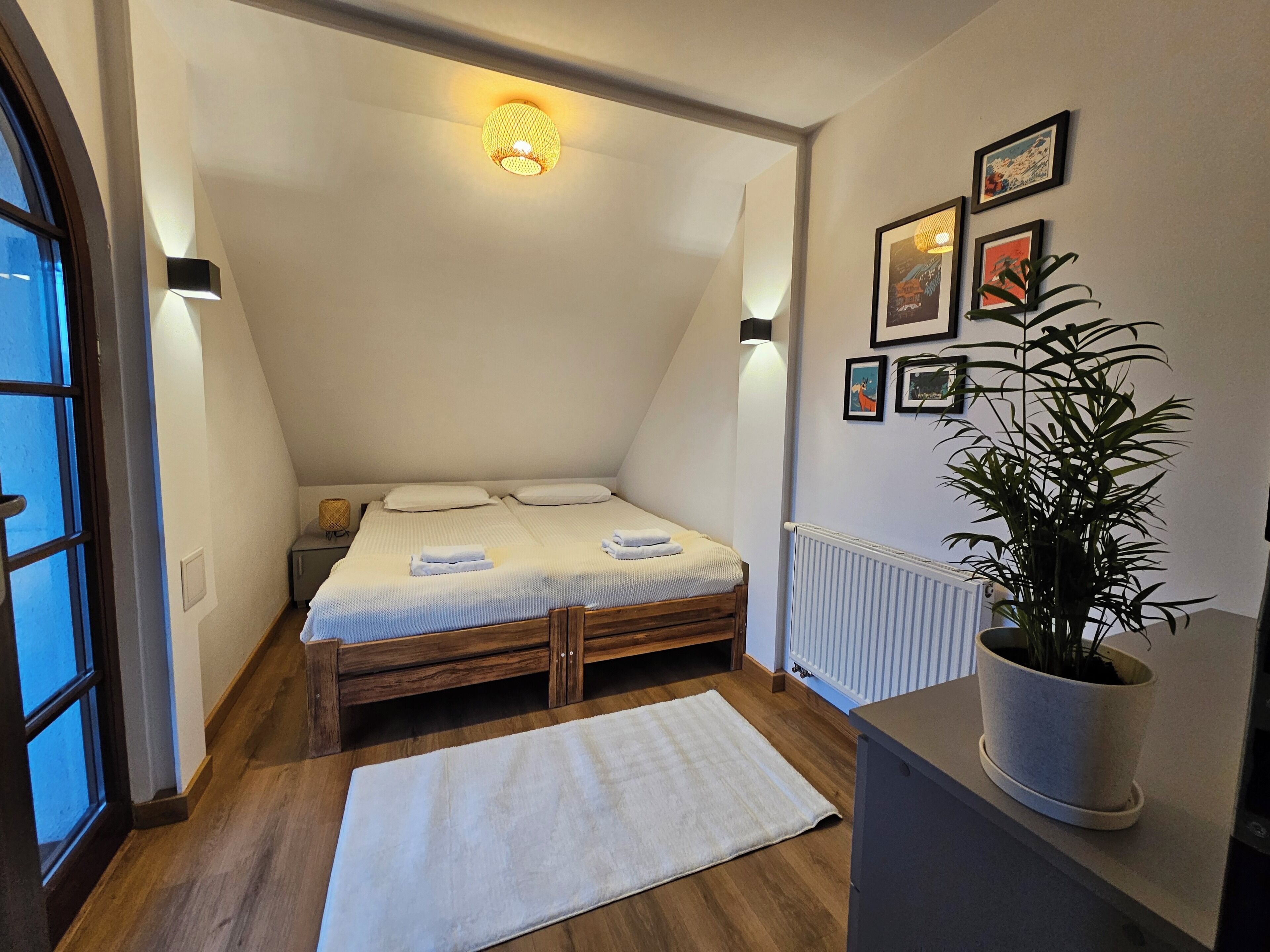 A small, cozy bedroom with a sloped ceiling, a wooden bed, a white rug, a potted plant, and framed pictures on the wall.