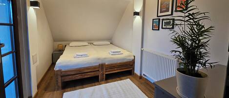A small, cozy bedroom with a sloped ceiling, a wooden bed, a white rug, a potted plant, and framed pictures on the wall.