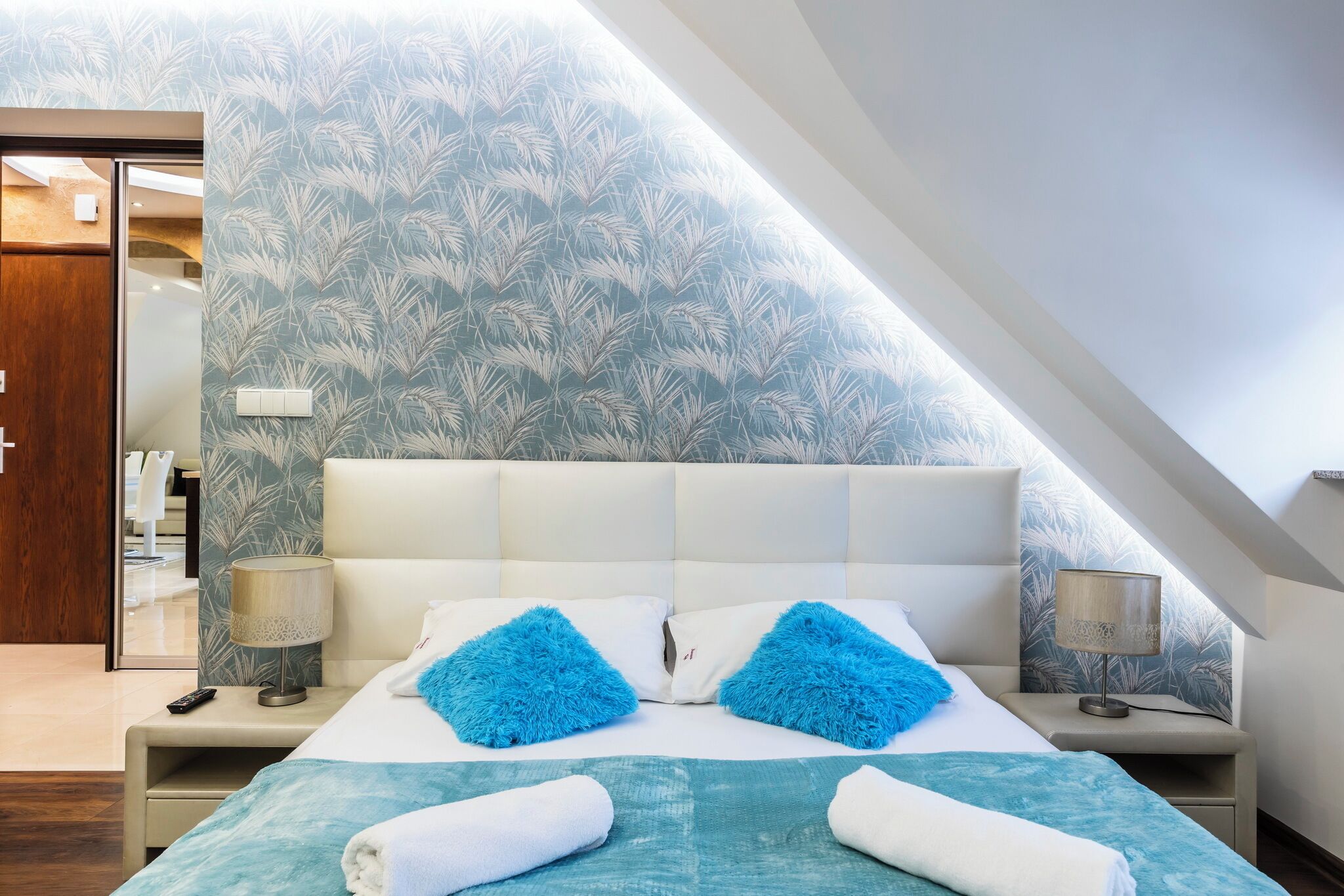 Una habitación de hotel moderna con una cama grande, dos almohadas mullidas, una mesita de noche con lámpara y un espejo.