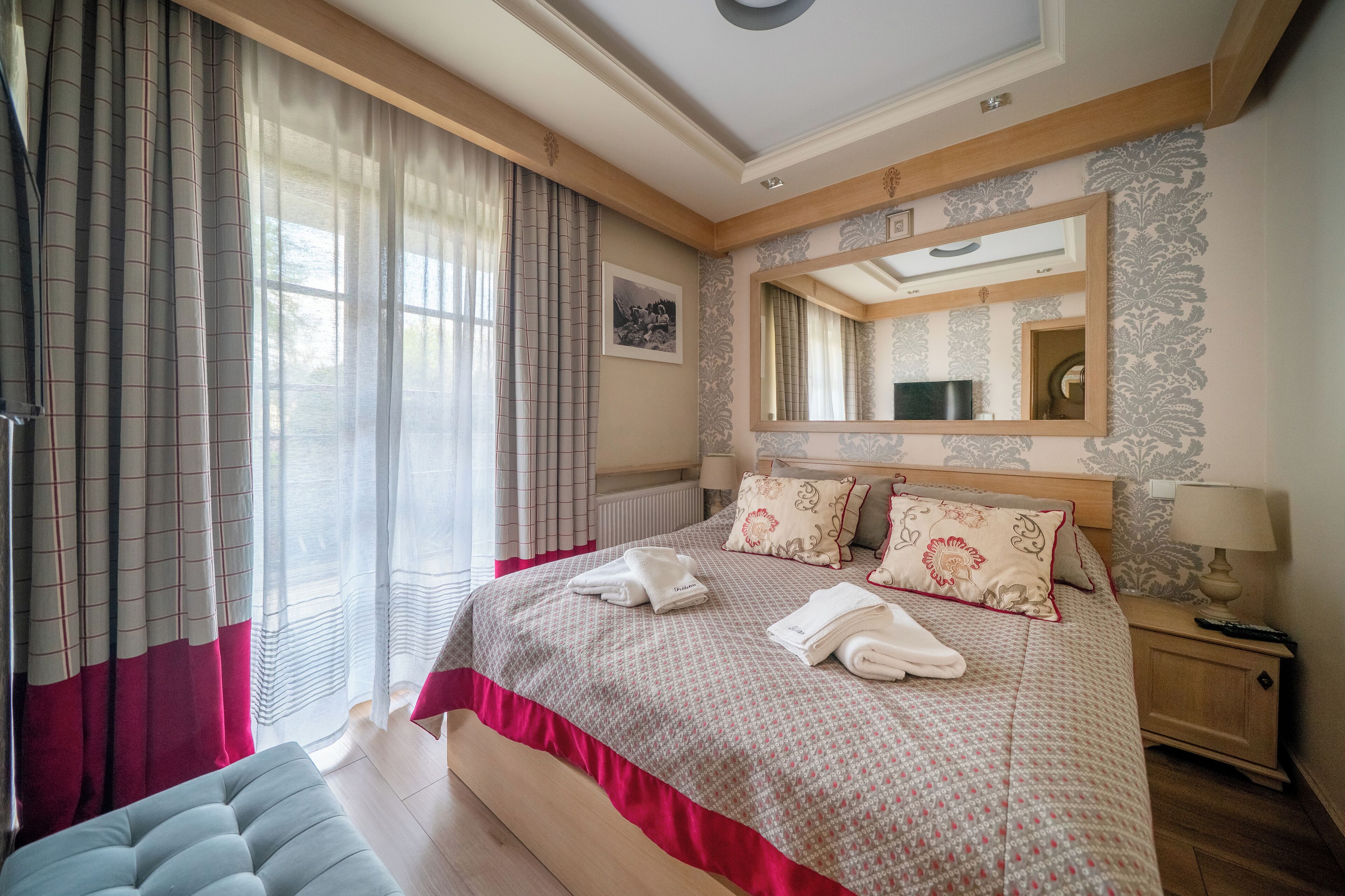 A well-lit bedroom with a large bed, a bedside table with a lamp, and a window with curtains.