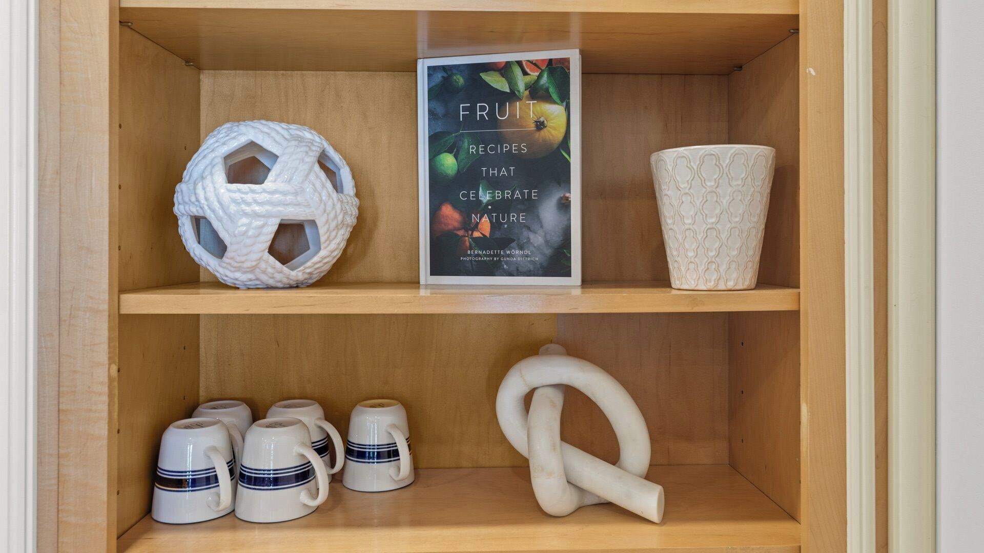 Una estantería de madera con un libro titulado 'Recetas de Frutas que Celebran la Naturaleza', una cesta tejida, una taza de cerámica y un juego de tazas.