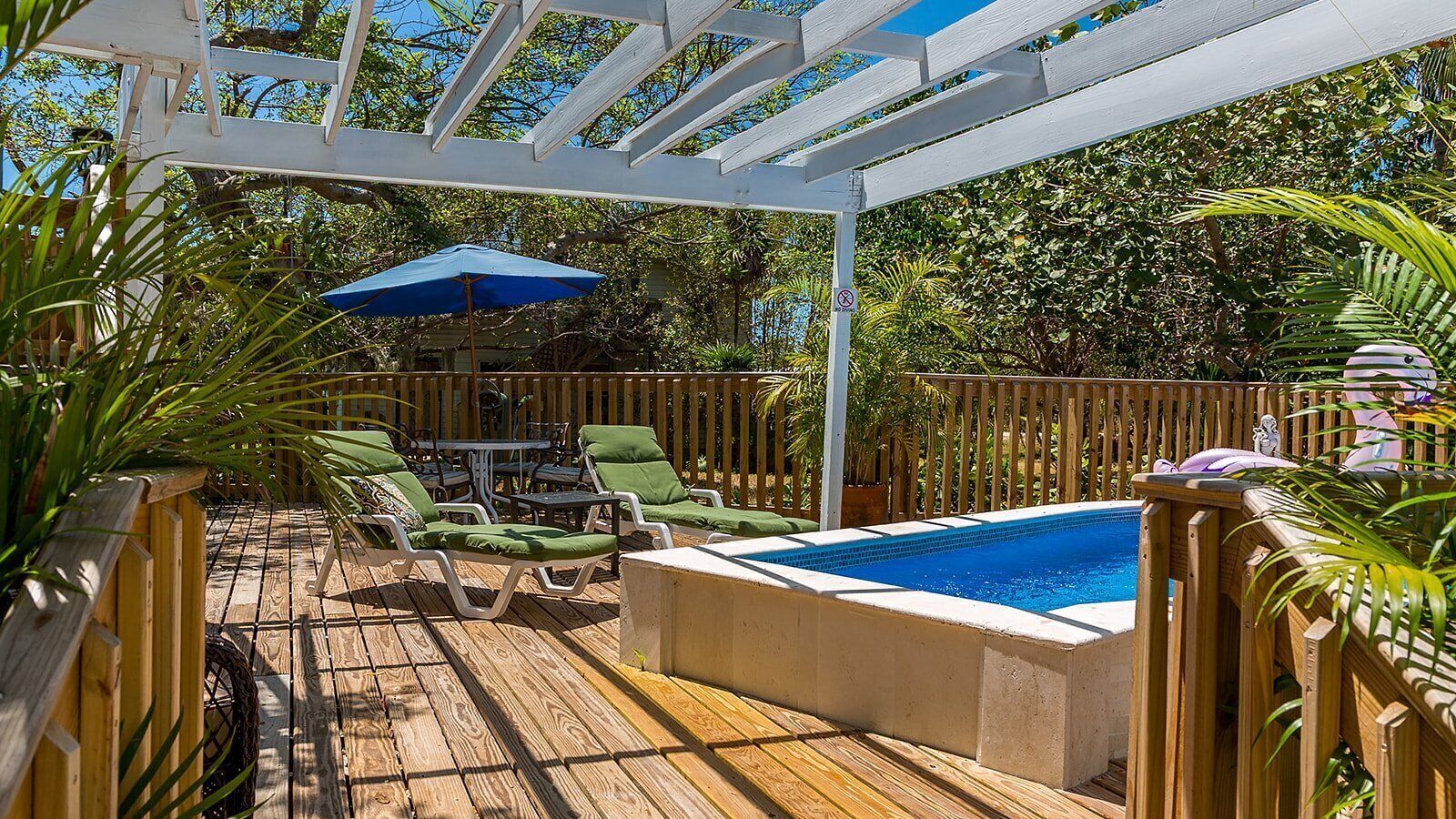 A wooden deck with a pool, lounge chairs, and a pergola with a blue umbrella.
