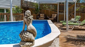 En swimmingpool med en havfruestatue, træterrasse, liggestole og et træhegn.
