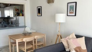 Una sala moderna con un sofá gris, una mesa de comedor de madera con sillas, una pequeña cocina y una lámpara de pie.