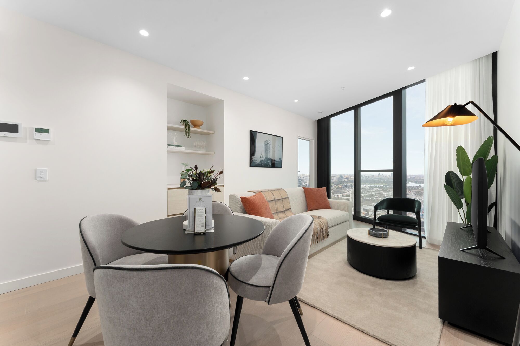 Een moderne woonkamer met een ronde tafel, twee grijze stoelen, een bank met kussens, een zwart bijzettafeltje en een groot raam met uitzicht op de stad.