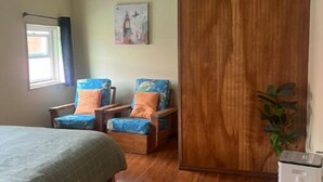 A room with a bed, two armchairs with cushions, a wooden wardrobe, a small white appliance, and a window with curtains. - Heart of Downtown Hilo (Hilo)