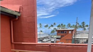 A wooden bench with a metal railing, a red wall, and a view of a street with parked cars and palm trees. - Heart of Downtown Hilo (Hilo)