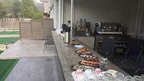 Un área de comedor al aire libre con una larga mesa dispuesta con diversos platos y bebidas, una cafetera y una parrilla.