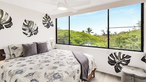 A bedroom with a large bed, a ceiling fan, and a view of lush greenery through a large window.