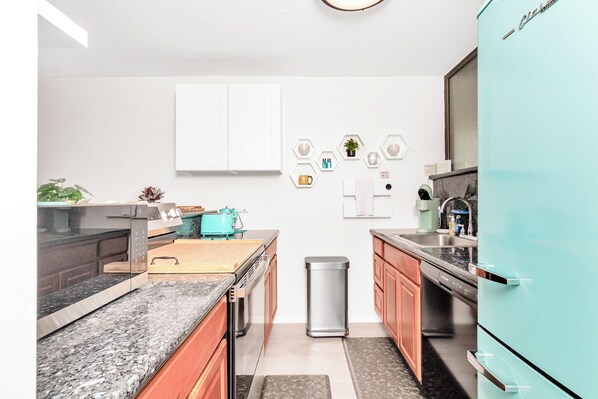 A modern kitchen with granite countertops, wooden cabinets, a teal refrigerator, and a small trash bin. - Ocean View Chic Condo at Carlsmith Beach Park in Hilo! (Hilo)