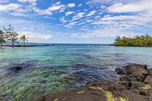 A clear view of a tropical beach with rocky shoreline, calm turquoise waters, and scattered palm trees. - Ocean View Chic Condo at Carlsmith Beach Park in Hilo! (Hilo)