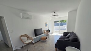 Ett modernt vardagsrum med en platt-TV, en mörk soffa, en fåtölj i rotting och ett litet vitt bord.